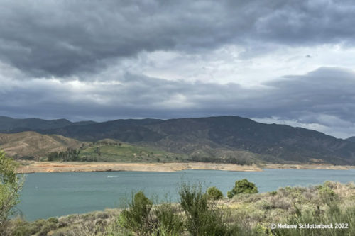 A lake with a bathtub ring showing lowered water levels against tall mountains in the back and storm clouds above.